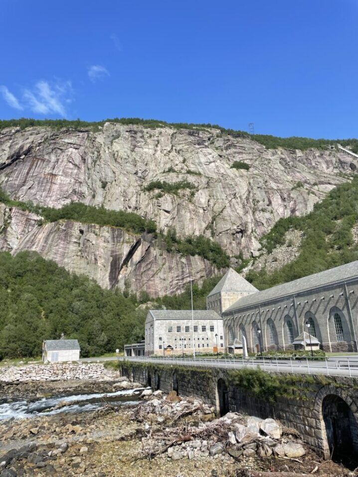 Kraftverket midt i bildet. Skog og fjell i bakgrunnen og en knall blå himmel øverst. En kan se en elv som renner ut under kraftverket.