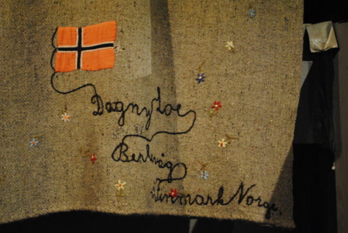 Broderi på lys, brunt teppe. Viser et norsk flagg øverst til venstre, noen små blomster spredt utover og så er det brodert med løkkeskrift: Dagny Loe, Berlevåg, Finnmark, Norge.