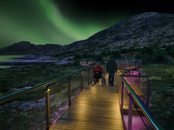 En opplyst trebro går gjennom et åpent naturområde om natten. To personer beveger seg langs broen, hvorav én bruker en rullestol. I bakgrunnen ligger fjellformasjoner og et svakt opplyst landskap, mens nordlyset sveiper over nattehimmelen.