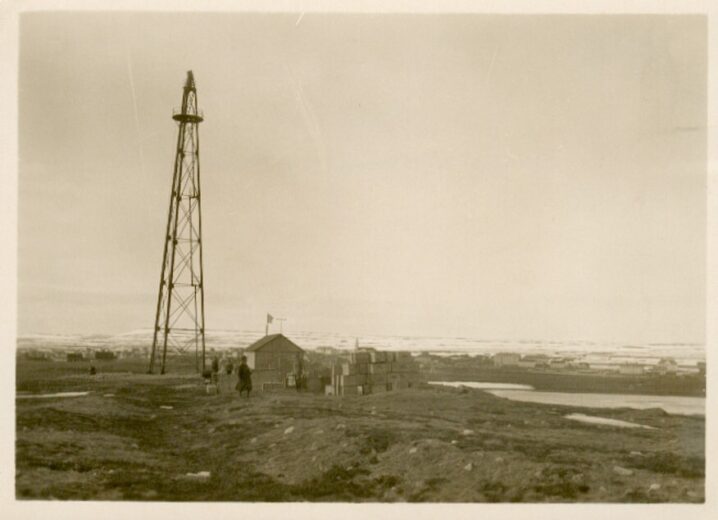 Svart hvitt bilde av luftskipmasta. Slette med et lite hus og noen mennesker foran.