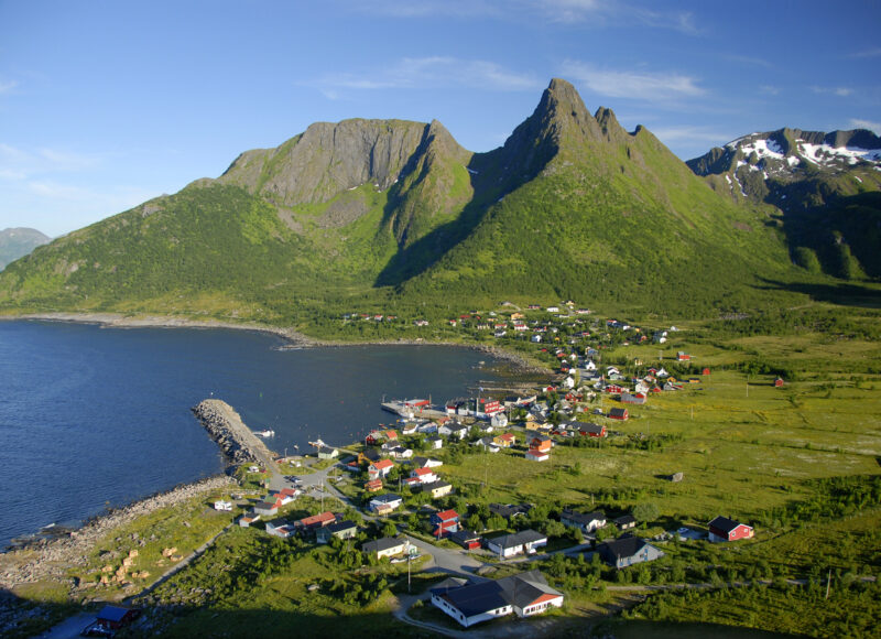 Fiskevær innerst i en liten fjord sett ovenfra. Spisse grønne fjell i bakgrunnen og knall blå himmel.
