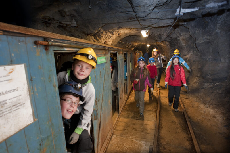 Flere barn inne i en gruve. Non går mot kamera og et par stikker ut fra et lavt, blåmalt bygg inne i gruva. Alle har på hjelm.