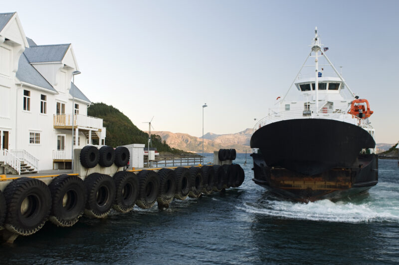 Stor bilferge kommer mot på vannet på vei mot kai. Kai dekket av bildekk. Stor hvit brygge på land. Fjell i bakgrunnen og blå himmel.
