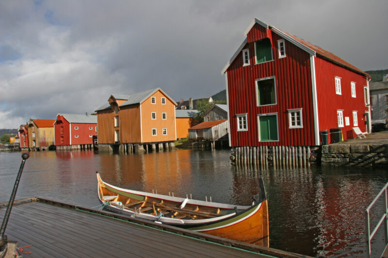 En nordlandsbåt ligger bed brygge på fjorden i forgrunnen. Bak dere en rekke med gamle brygger i rødt og oker.