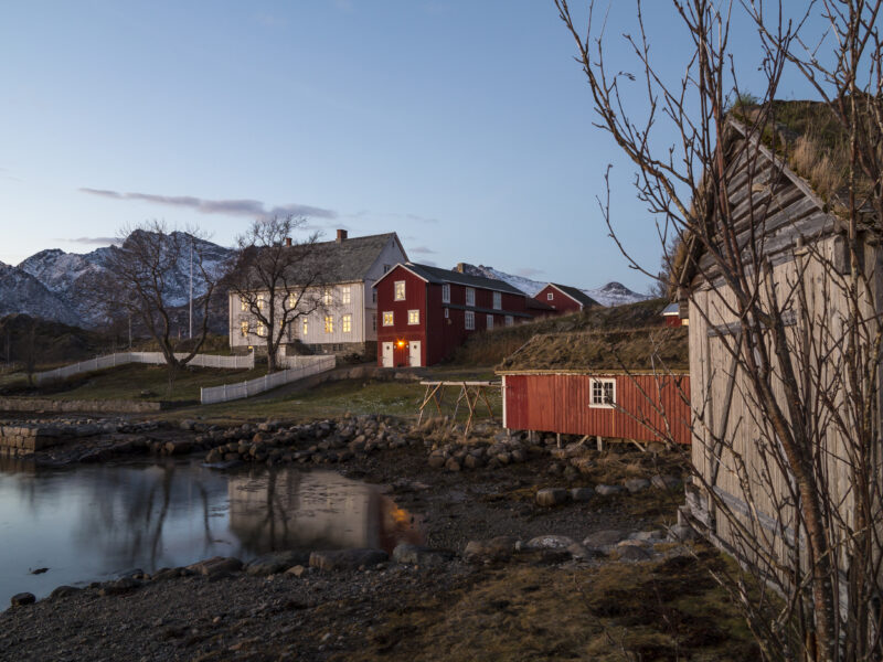 Bildet viser et rolig landskap ved vannkanten i skumringen. Flere hus i røde og lyse farger ligger spredt langs bredden, med fjell som reiser seg i bakgrunnen. Vannet i forgrunnen speiler husene og den dempede himmelen, og trær uten blader står rundt bebyggelsen.