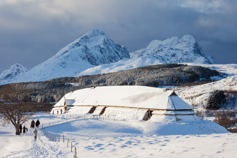 Bildet viser et snødekt vinterlandskap med et snødekt langhus omgitt av gjerder i forgrunnen. To personer går langs en brøytet vei i snøen. I bakgrunnen ruver høye, snødekte fjell under en overskyet himmel.