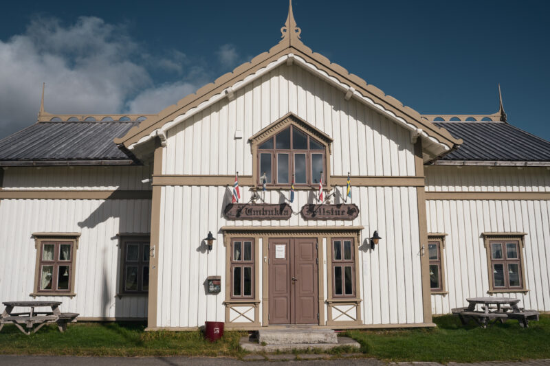 Hvitmalt fjellstue med lys, brune kanter. Blå himmel med noen skyer over.