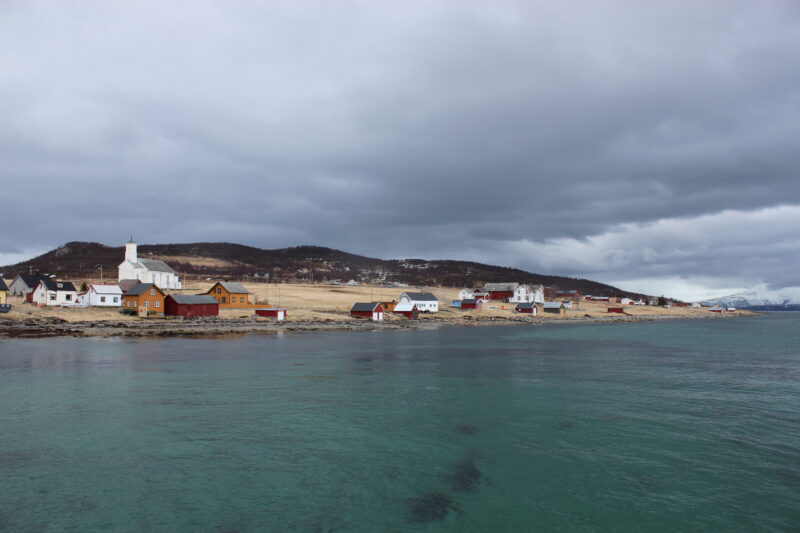 Et kystlandskap med klart, turkist hav i forgrunnen og en rekke spredte bygninger langs strandlinjen. Husene er hovedsakelig lave trebygninger i lyse og røde farger. Landskapet bak består av gress- og lyngkledde åser under en overskyet, grå himmel.