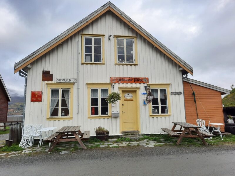 "Et hvitt, toetasjes trehus med gult panel og røde detaljer, omgitt av benker og en grusvei, med fjell i bakgrunnen under en overskyet himmel."