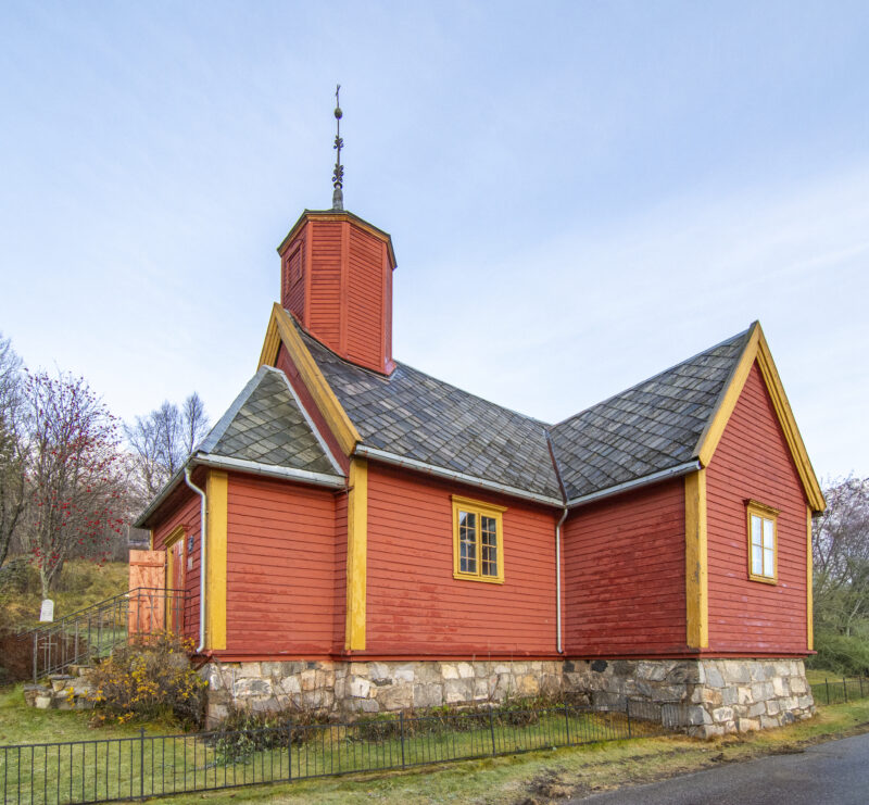 En liten, rød trekirke med gule hjørnelister og steinsokkel, omgitt av gress og trær. Bygget har en lav tårnkonstruksjon med spir og skifertekket tak. Himmelel er lys og klar.