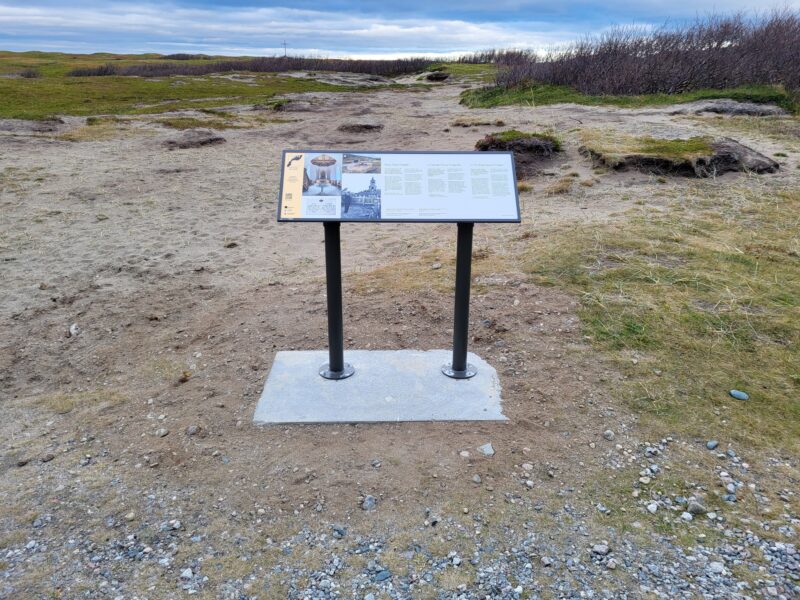 "Et informasjonsskilt på en ståramme i et åpent, karrig landskap med gras og fjell i bakgrunnen."