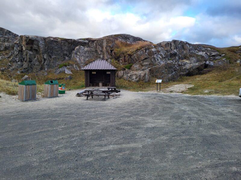 «Et friluftsområde med en åpen, gruslagt plass omgitt av fjell og lav vegetasjon. Midt i bildet står et lite overbygd gapahuk‑lignende bygg med sitteplasser, og foran det står et piknikbord. Til venstre står tre avfallsbeholdere, og til høyre sees en informasjonstavle langs stien. Himmelen er overskyet.»