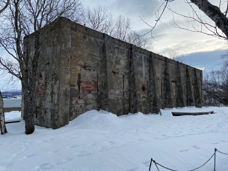 En ruin av et industribygg. Foran noen tre og snø på bakken. Blå himmel med skyer over.