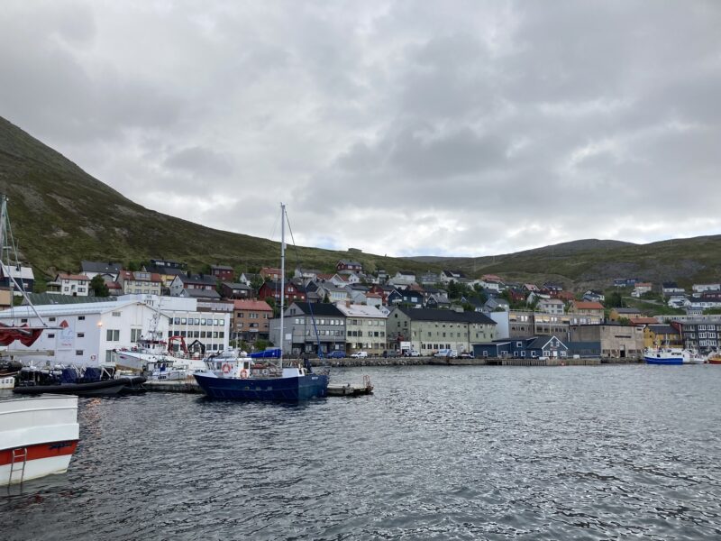 Bilde av Honningsvåg fra sjøen. Sjø i forgrunnen, noen båter og så tett bebyggelse. En ås i bakgrunnen og en himmel med tett skydekke.