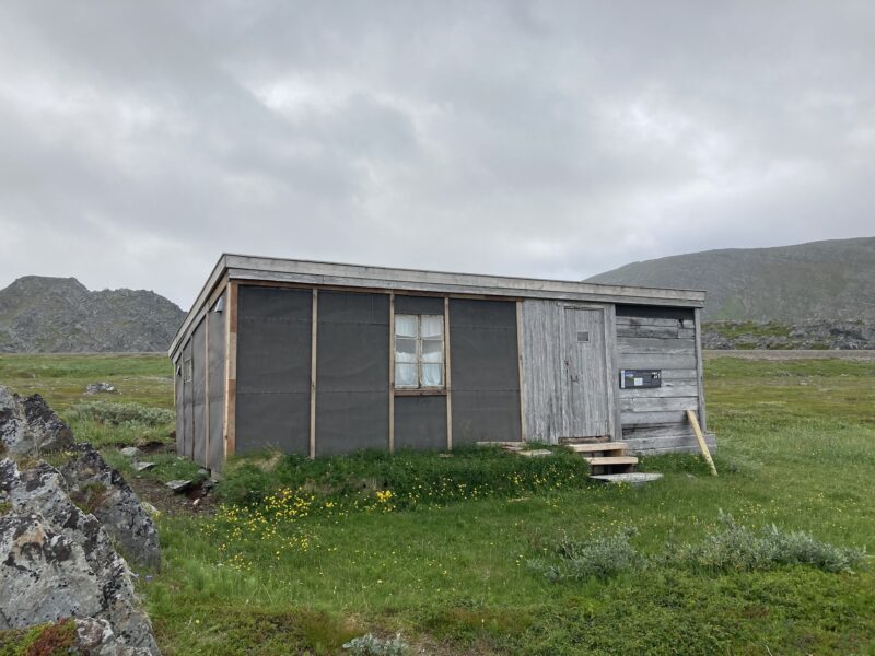 Enkel hytte under rekonstruksjon i midten av bildet. Gress på bakken rundt. Fjell i bakgrunnen og himmel med tett skydekke.
