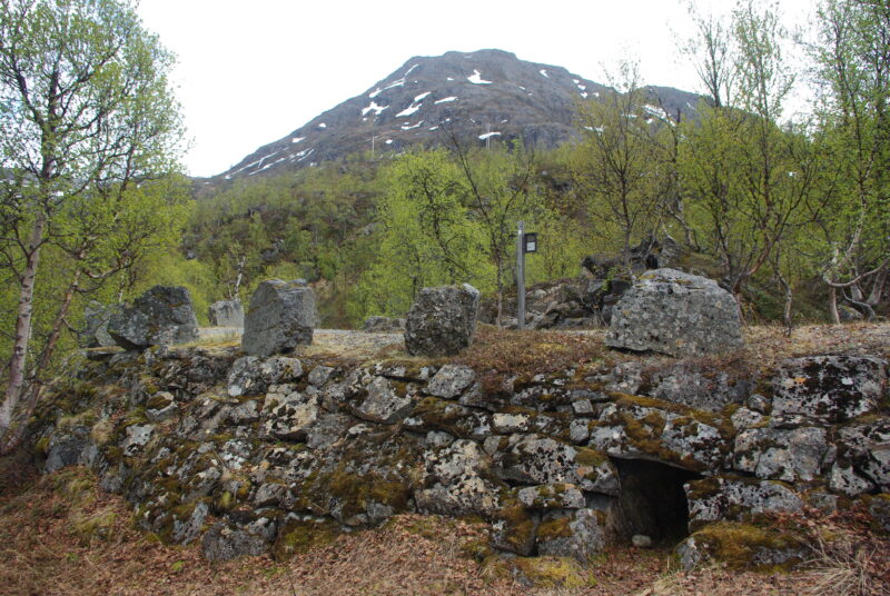 Gammel mur ved vei, større steiner langs veien over. Grusvei skimtes over muren og bak der noe skog, en fjelltopp og hvit himmel.
