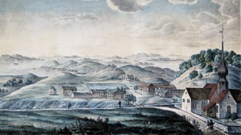 Gammelt maleri av kirkested med kirke til høyre og en rekke andre bygninger til venstre. Landskap bak med åser, fjell og hav.