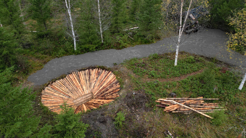 Ser ned på rekonstruert fangstgrop med tømmer. Grusvei bak og noen tømmerstokker til høyre. Vegetasjon og noen trær rundt.