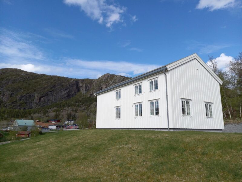 Liten bakketopp med grønt gress. På toppen et hvitt, gammelt hus. Til venstre, litt lengre bort, noen bygninger. Over dem en ås. Blå himmel med noen få hvite skyer.