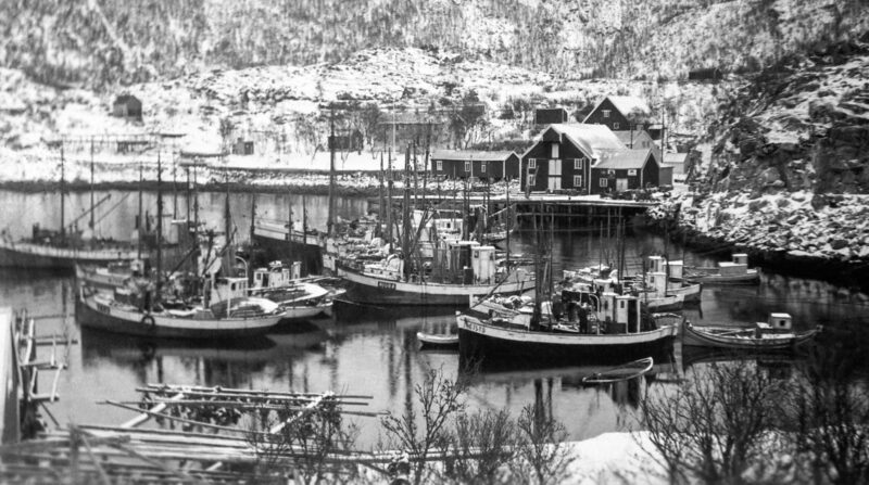 Svart‑hvitt fotografi av en liten havn med flere fiskebåter fortøyd ved brygger. Rundt havnen ligger snødekte bygninger og terreng, omkranset av vinterkledde fjellsider.