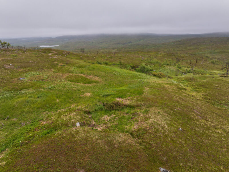 Et grønnkledt landskap med en forhøyning i forgrunnen og spor etter bosetninger. Viddelandskap i bakgrunnen og en himmel med tett skydekke.