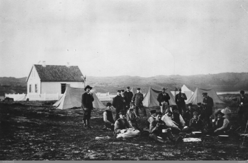 Svart hvitt bilde. Arbeidere står og sitter på en vidde foran noen telt. Hvitt hus i bakgrunnen. Fjell lengst bak.