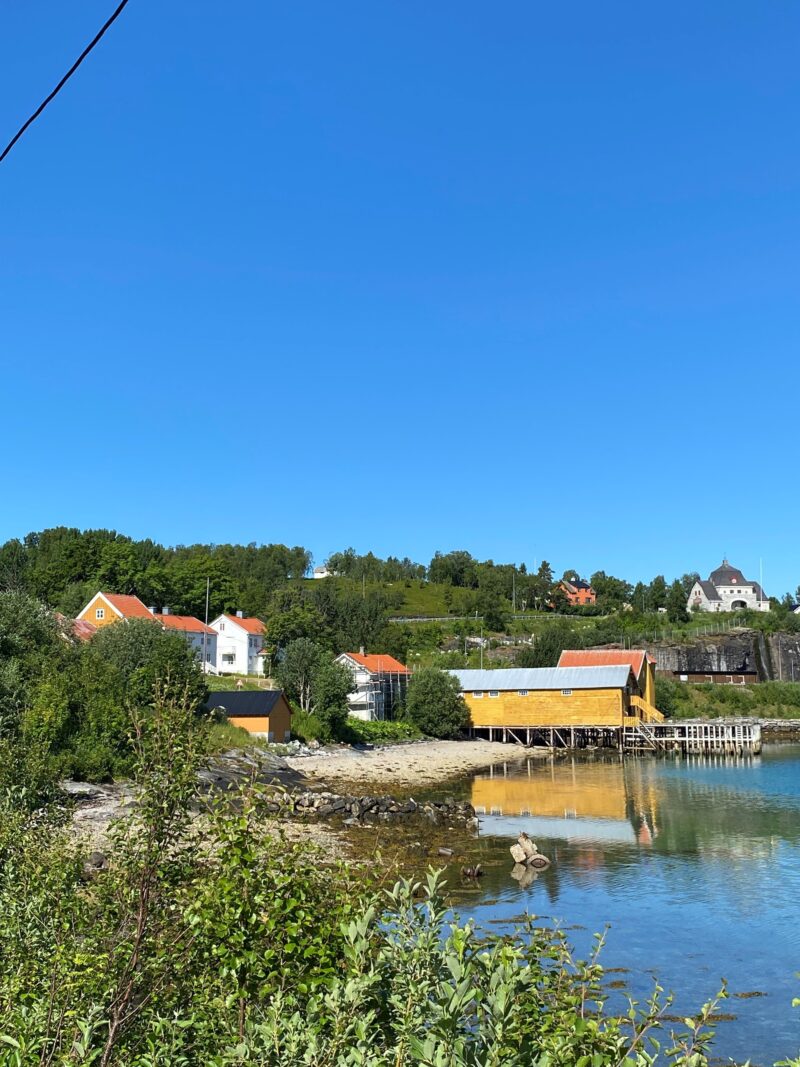 Sommerlig kystlandskap med klart blå himmel. Flere gule og hvite trehus ligger langs vannkanten, med grønne trær og busker rundt. Vannet er stille og speiler bygningene og terrenget. Bakenfor ligger et lite kulturlandskap med spredte hus og gresskledde bakker.
