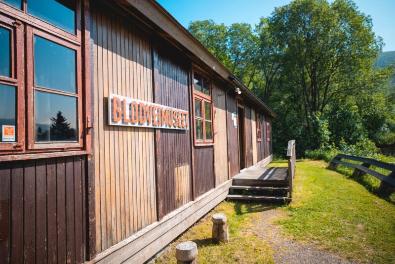"Brunt trebygg med vinduer og inngangsparti langs en grussti. Bygningen ligger i grønne og solrike omgivelser med trær og gjerde i bakgrunnen. Et skilt er montert på veggen."