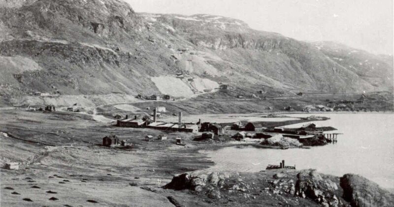 Historisk svart‑hvitt‑foto av et lite industristed ved en fjord, omgitt av fjell. Flere lave bygninger, fabrikkpiper og bryggeanlegg ligger spredt langs vannkanten. Landskapet rundt består av åpne vidder og nakne fjellsider.
