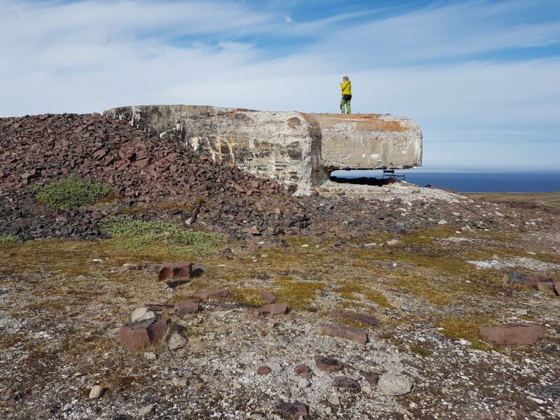 «En åpen, steinete vidde med lav vegetasjon og ujevnt terreng. Midt i bildet ligger en stor, rektangulær betongstruktur som ser ut som en eldre bunkerlignende konstruksjon. En person i en gul jakke står oppe på konstruksjonen og skuer utover området. I bakgrunnen er himmelen lys og delvis skyet, med antydning til hav i horisonten.»