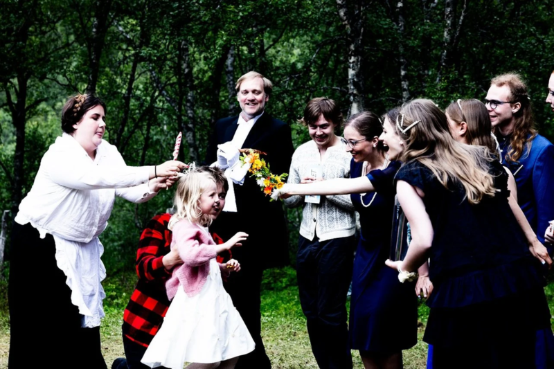 "En gruppe mennesker samles utendørs i en skogkledd omgivelse, der to kvinner i hvite kjoler og en jente i rosa kjole holder en blomsterdekket stang eller sløyfe, omgitt av smilende tilskuere."