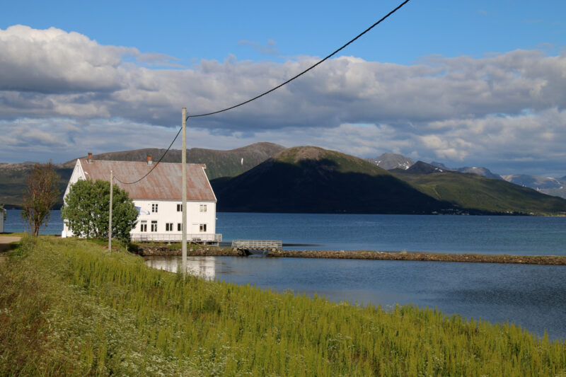 En stor hvit brygge i midten/til venstre i bildet. Gress forrest i bildet. Brygga ligger ved vannet. Fjell i bakgrunnen. Blå himmel med skyer over.