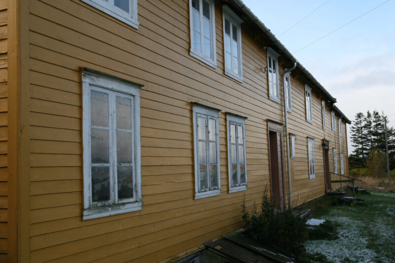 Gammelt, gulmalt hus. Noe gress foran til høyre. Noen trær og blå himmel bak.