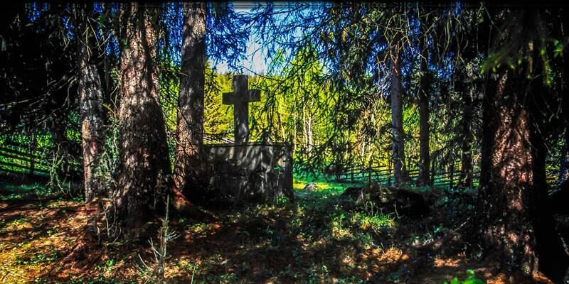 Et skogsområde med høye trær som danner skygge rundt et gammelt steinmonument med et kors. Sollys slipper inn mellom trærne og lyser opp gresset i bakgrunnen.