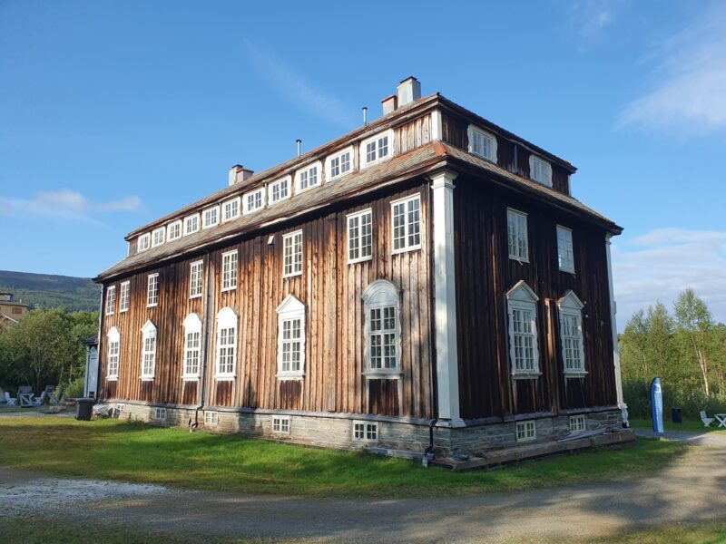 Stort gammel bygg i tre. Malt brunt med hvite karmer. Gress og grus rundt bygget. En ås i bakgrunnen og blå himmel.