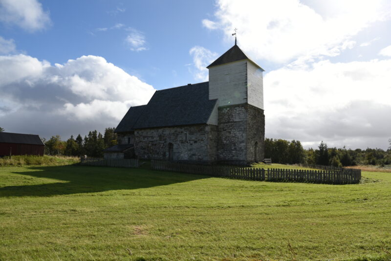 Stor gresslette i forgrunnen og så en gammel, hvit og grå steinkirke i midten. Noen trær bak. Blå himmel med skyer.