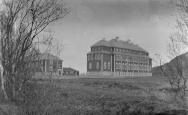 Gammelt svart‑hvitt‑fotografi som viser to store murbygninger i et åpent landskap. Bygningene har flere etasjer, symmetriske vindusrekker og bratte tak. Trær uten løv rammer inn motivet i forgrunnen, og bakken foran bygningene er gresskledd og ujevn.