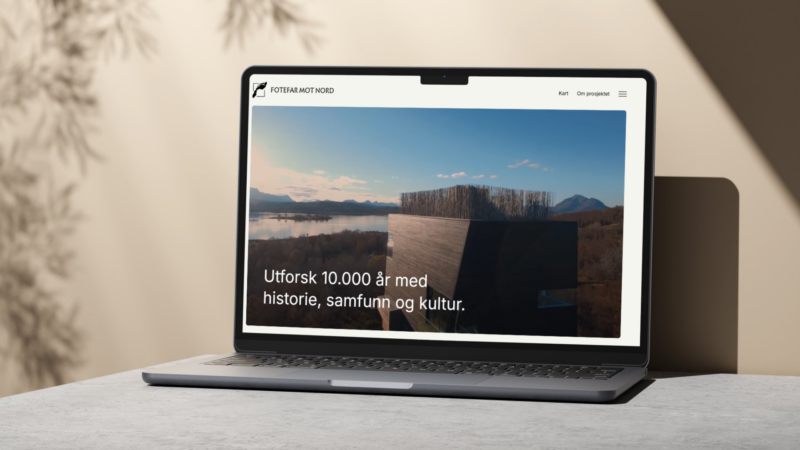 "En bærbar datamaskin som viser nettsiden til Fotefar Mot Nord. På skjermen er det et bilde av et moderne bygningselement med utsikt over et landskap med fjell, vann og skog. Over bildet står teksten: 'Utforsk 10.000 år med historie, samfunn og kultur.' Nettsiden har en navigasjonsmeny øverst med lenker som 'Kart' og 'Om prosjektet'."