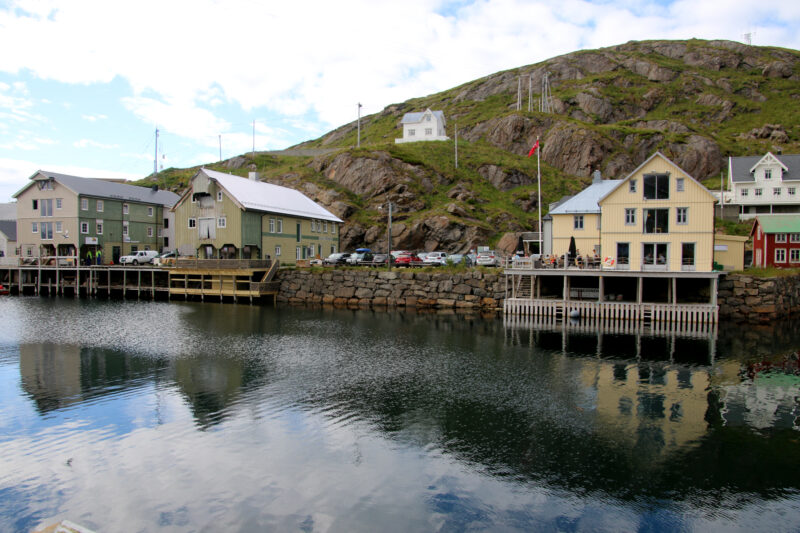 Noen gamle bygninger i et nedlagt fiskevær sett fra sjøen. En lien gresskledt høyde bak et et lite hvitt hus. Blå himmel med skyer over.