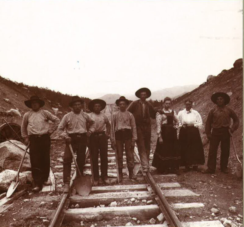 Svart hvitt bilde. Menn og kvinner med hakker og annet utstyr står på jernbaneskinner. Steiner og grus rundt. Fjell i bakgrunnen. Hvit himmel.