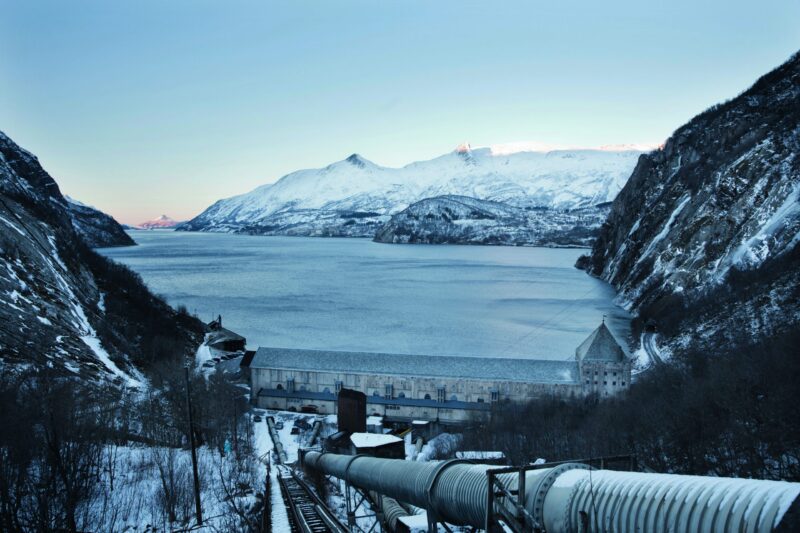 Kraftverk i forgrunnen med et stort rør som går opp mot fotografen. Nederst og innerst i en fjord ligger kraftverket. Utsikt over fjorden og mot snødekte fjell.