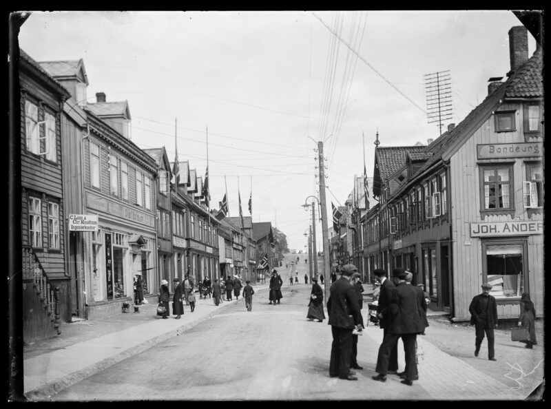Svart hvitt bilde. Bygate med hus på begge sider. Flere mennesker går i gaten. Hvit himmel over.