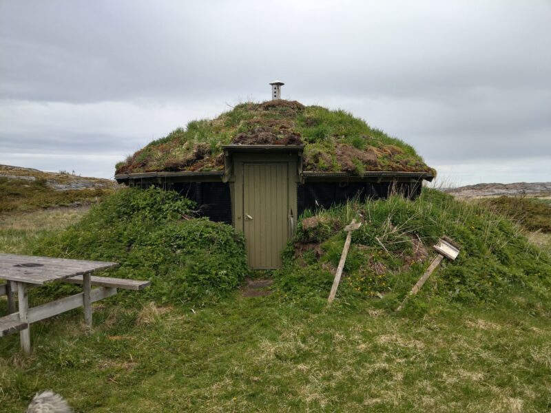 En liten, rund bygning med torvtak og en liten dør. Gress rundt. En benk i tre til høyre og et par skilt som nesten har falt ned. Himmel med tett skydekke over.