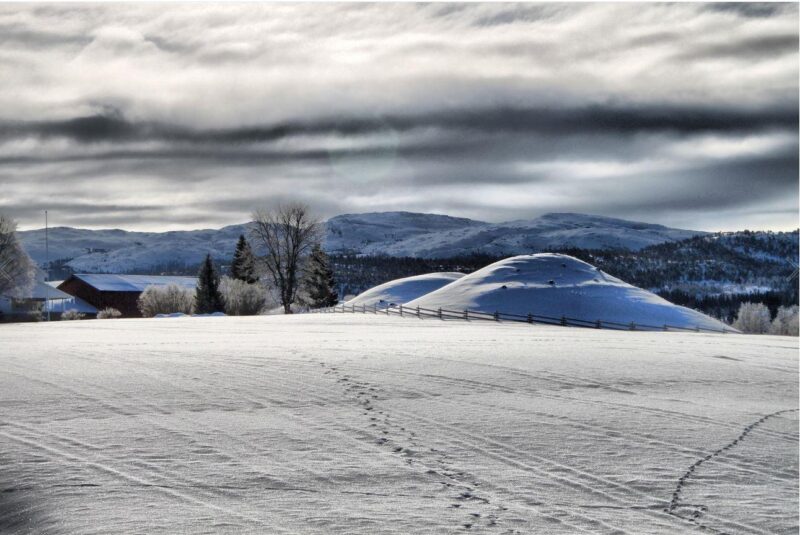Snødekt slette og to gravhauger i enden, også dekket av snø. Bak der utsikt mot landskap med trær, neon få bygninger, fjell og himmel med skyer.