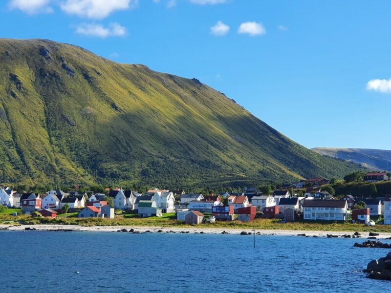 En landsby sett fra havet. Hav foran, en klynge med hus, et fjell kledt i grønt bak og så blå himmel.