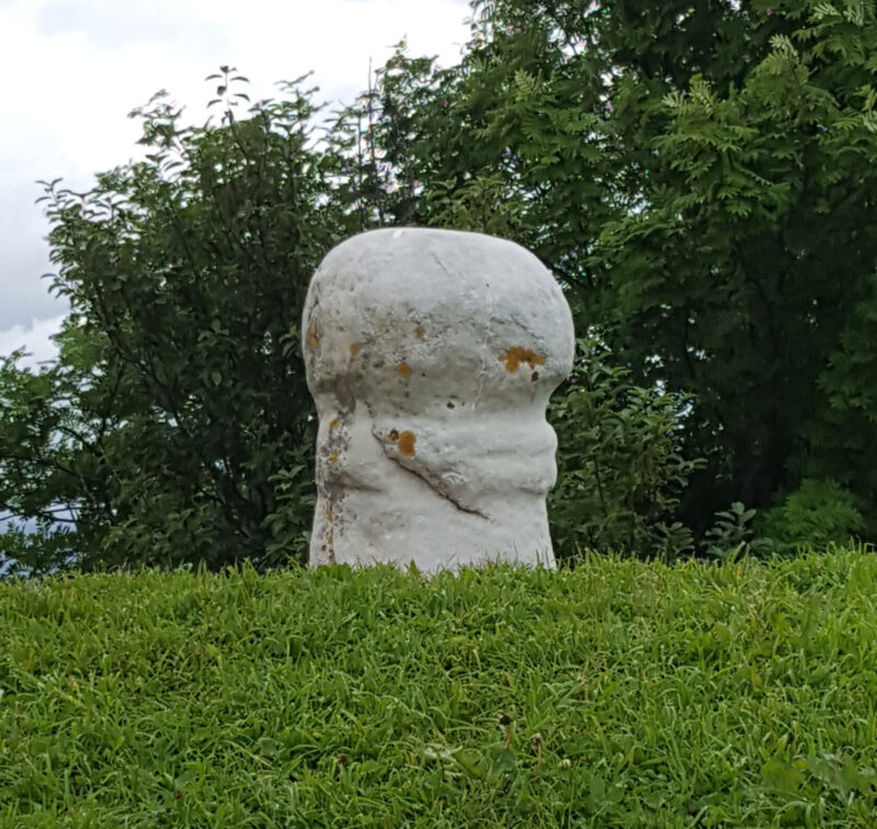 Liten, lys steinskulptur med avrundet form står på en gresskledd bakke, omgitt av trær og under en overskyet himmel.