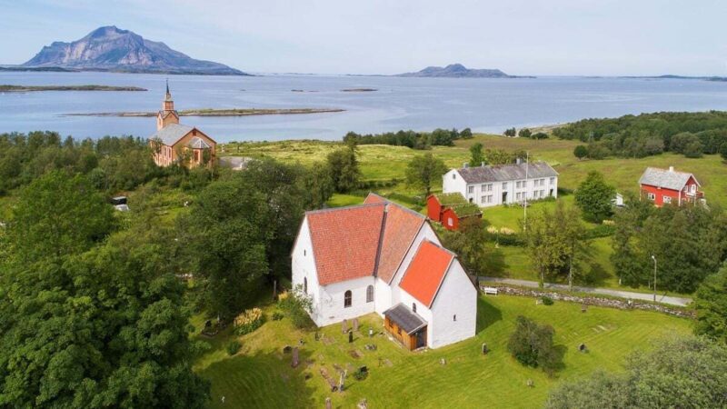Flyfoto av et grønt kystlandskap med flere bygninger, inkludert en hvit kirke med rødt tak i forgrunnen og en større steinkirke lenger bak. Rundt bygningene ligger åpne jorder, trær og spredte hus. I bakgrunnen skimtes havet og flere fjellformasjoner.