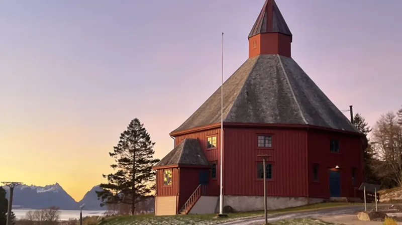 En rød, gammel, sekskantet kirke med grått tak til høyre i bildet. Noen trær rundt. Fjell i bakgrunnen og en himmel med ulike farger som viser solnedgang.