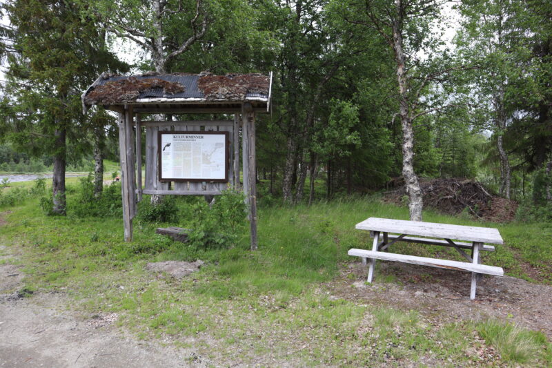 En grusvei forrest og så noe gress. Det står en trebenk til høyre og et stort skilt med treramme og tak til venstre. Skog bak.