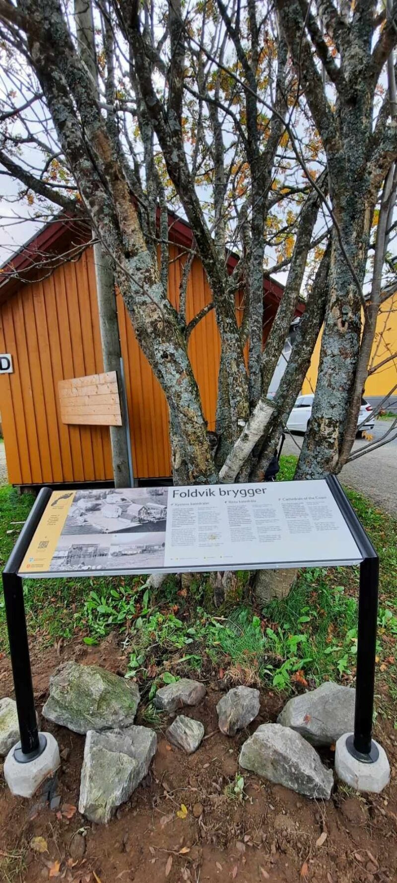 Stort informasjonskilt foran tre og en orangemalt trebrygge